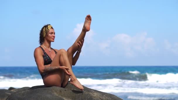 Mujer haciendo ejercicio en la playa con arena negra volcánica en un traje de baño sentado en una roca alrededor de las montañas de lava. El concepto de estilo de vida saludable y recreación en las Islas — Vídeo de stock