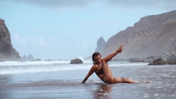 Una donna impegnata in ginnastica sull'oceano siede sullo spago ed esegue un ponte vicino all'acqua contro le onde e. Pratica di yoga. Uno stile di vita sano. Allungamento — Video Stock