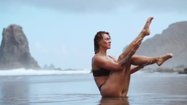 Sportlerin trainiert Bauchmuskeln am Strand mit schwarzem Sand vor Felsen und Wellen — Stockvideo