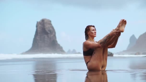 Femme sportive Entraînement des muscles abdominaux sur la plage avec du sable noir sur fond de rochers et de vagues — Video