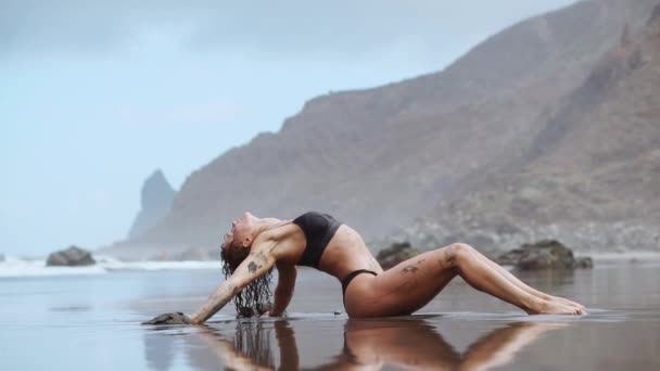 Vrouw beweegt plastisch dansen op het zand in de buurt van de Oceaan tegen de bergen en zwart zand — Stockvideo