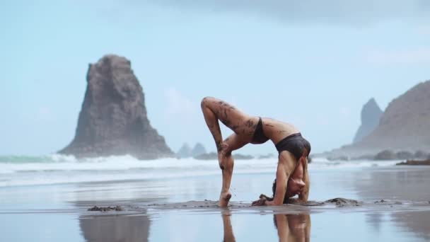 Uma bela mulher de fato de banho realiza uma ponte de exercício na praia com areia vulcânica preta rodeada por montanhas e oceano. Ginástica e ioga — Vídeo de Stock