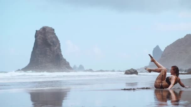 Donna si muove plasticamente ballando sulla sabbia vicino all'oceano contro le montagne e la sabbia nera — Video Stock