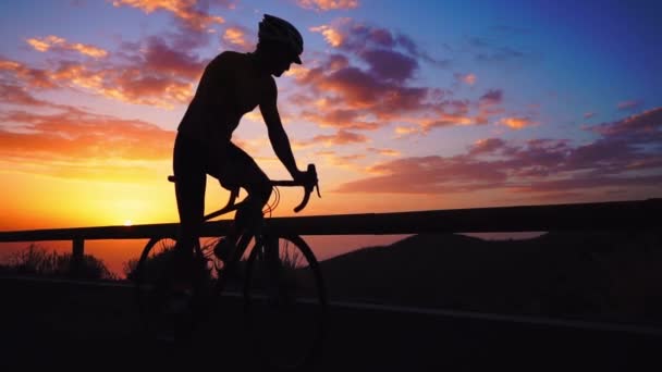 Silhuetten av en man som rider en cykel vid solnedgången på väg sida bergsutsikt. Slow motion steadicam — Stockvideo