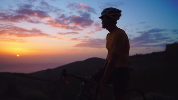 Молодий спортивний чоловік сидить на велосипеді на вершині гори в жовтому футбольному шоломі та спортивному обладнанні, дивлячись на захід сонця. Повільна бігова доріжка руху — стокове відео