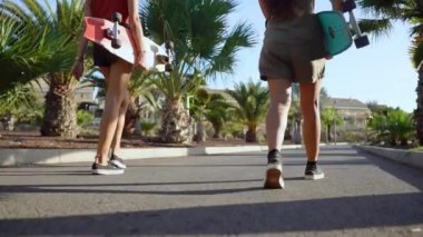 İki genç kadın longboards ve Kolo palmiye ağaçlarının ayar güneş altında elinde ile parkta yürümek. Ağır çekimde arkadan görünüm