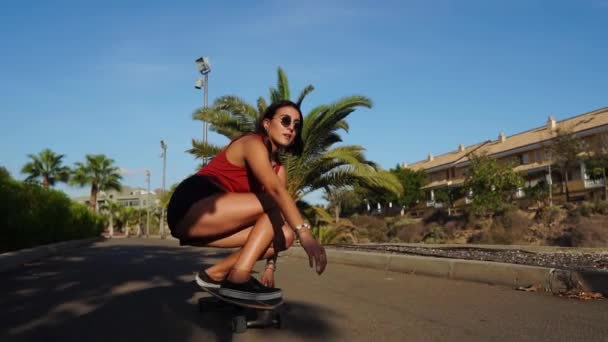 Молодая женщина летом катается на длинной доске возле пальм в шортах и кроссовках — стоковое видео