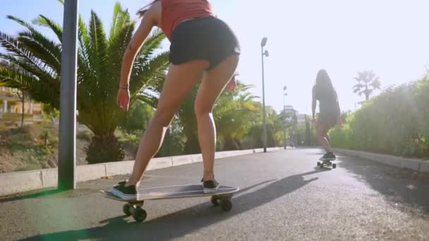 Two girls in skate Park ride along the track in sunlight on longboards looking forward and laughing in slow motion. steadicam back view — Stock Video