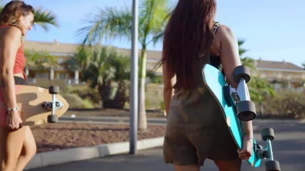 Dos chicas caminando al atardecer sonriendo con tablas para patinar Junta a lo largo del camino en el Parque con palmeras y arena — Vídeo de stock