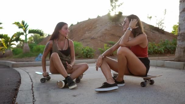 スケート公園で二人の女の子はボードの上に座るし、微笑し、夕日の光の中の冗談に笑って話します。ロングボード会話友達 — ストック動画