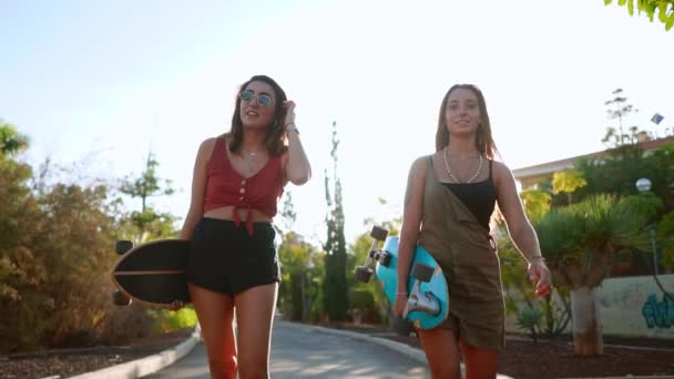 Девушки идут в парк, держа скейтборды в руках, разговаривая и смеясь, улыбаясь друг другу на закате солнца в замедленной съемке. — стоковое видео