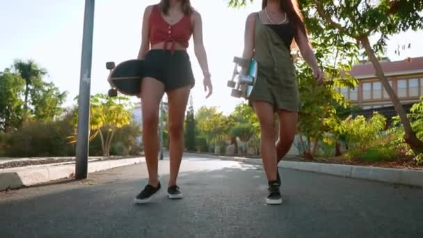 Девушки идут в парк, держа скейтборды в руках, разговаривая и смеясь, улыбаясь друг другу на закате солнца в замедленной съемке. — стоковое видео