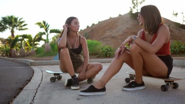 2 つのガール フレンドの笑いはスケート ボードの上に座って公園通信します。 — ストック動画