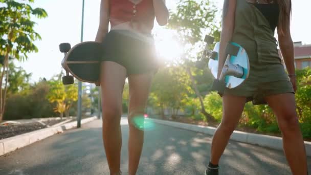Přítelkyně dívky chodí do parku drží skateboardy v jejich rukou, mluví a smáli se, usmívat se na sebe v západu slunce v pomalém pohybu — Stock video