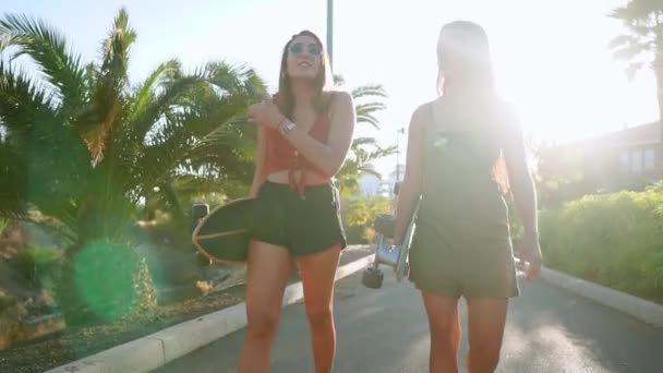 Chicas novias van al parque cerca de palmeras llevan patinetas en sus manos y hablando y riendo, sonriéndose entre sí en el sol del atardecer en cámara lenta — Vídeos de Stock