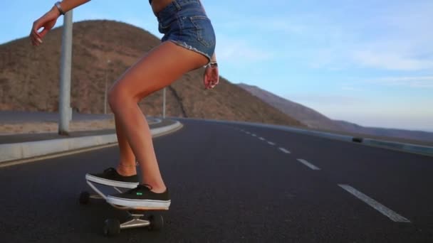 Дівчина в шортах і кросівках скейтбординг по дорозі на тлі океану і гір . — стокове відео
