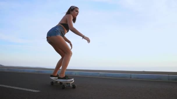 Chica en pantalones cortos y zapatillas de skate a lo largo de la carretera en el fondo del océano y las montañas . — Vídeos de Stock