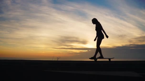 Silhouette eines Mädchens in kurzen Hosen und Turnschuhen, das auf der Straße gegen Meer und Berge skateboardet. — Stockvideo