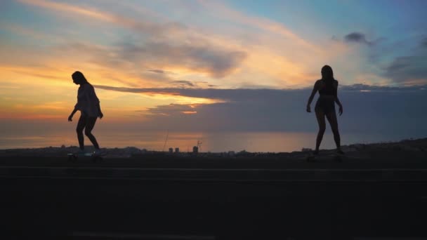Силуэт двух девушек, катающихся на скейтбордах на фоне океана и закатного неба — стоковое видео