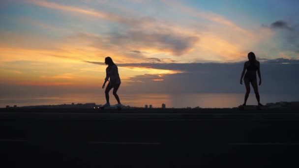 Silueta de dos chicas montando monopatines en el fondo del océano y el cielo puesta de sol — Vídeo de stock