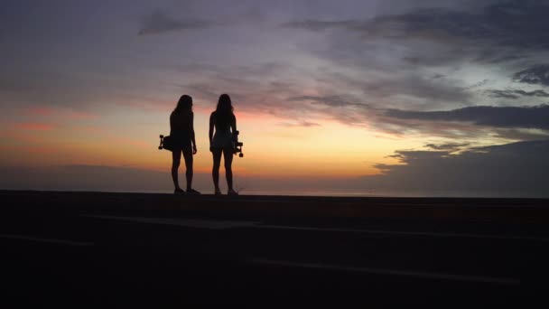 Two girls standing on the edge of the road with skateboards in their hands watching the sunset — Wideo stockowe