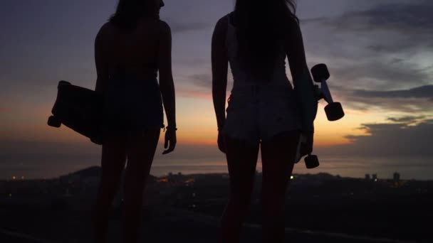 Две девушки стоят на краю дороги со скейтбордами в руках и смотрят на закат — стоковое видео
