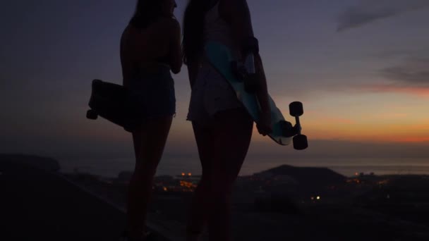 Two girls standing on the edge of the road with skateboards in their hands watching the sunset — Wideo stockowe