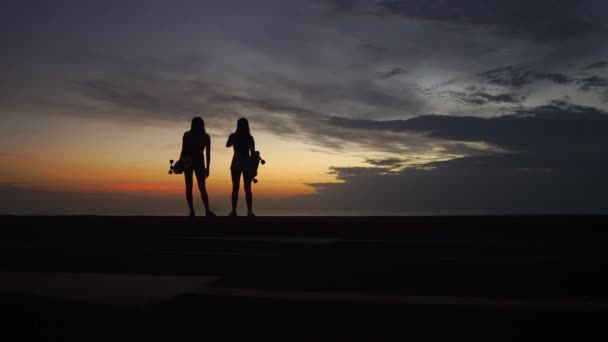Дві дівчини стоять на краю дороги зі скейтбордами в руках, спостерігаючи за заходом сонця — стокове відео