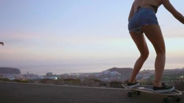 Kız skeytbordistka gitmek için gülümseyen ve okyanus ve yavaşlatılmış dağlarda arka plan üzerinde gülüyor dağlar ateş kısa şort gün batımında