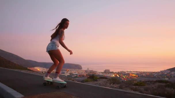 Красивая девушка катается на скейтборде по дороге против закатного неба — стоковое видео