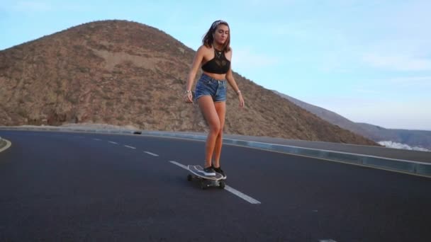 Chica en pantalones cortos y zapatillas de skate a lo largo de la carretera en el fondo del océano y las montañas . — Vídeos de Stock