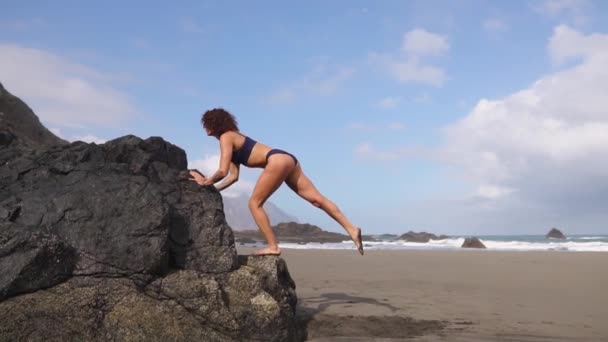 Esbelta zhenina en la playa con arena volcánica dedicada a los deportes en un lugar muy hermoso haciendo sentadillas en su pierna apoyada en una piedra — Vídeo de stock