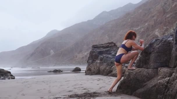 Atleta ragazza si esibisce squat su una gamba in piedi su una roccia sulla spiaggia con sabbia nera durante l'allenamento su una bella spiaggia. Stile di vita sano durante il viaggio — Video Stock