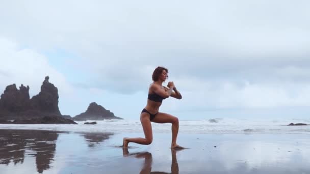 Вдоль океана, женщина занимается спортом выполняя выпады по очереди на каждой ноге на фоне камней и воды — стоковое видео