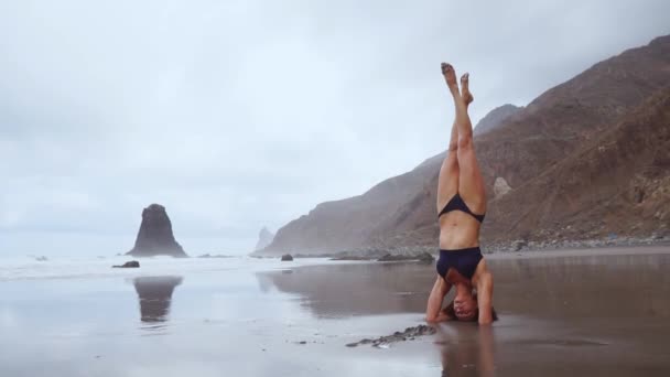 A lo largo del océano una mujer en cabeza de yoga y meditando. Estilo de vida saludable. Equilibrio en la cabeza de pie en la arena cerca del agua — Vídeo de stock