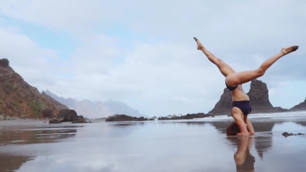 Chica de los deportes de pie sobre su cabeza en el océano mueve sus piernas realizando cordel en el aire. estiramiento — Vídeo de stock