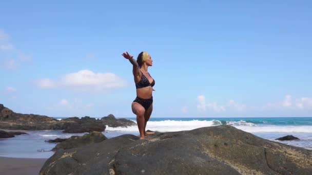 Mujer en armonía con la naturaleza medita de pie sobre una piedra en la playa cerca del océano — Vídeos de Stock