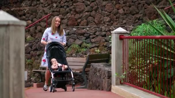 Eine junge Mutter geht mit einem Baby im Rollstuhl durch den Park mit Palmen und Wasserfall, eine glückliche Familie auf Reisen. — Stockvideo