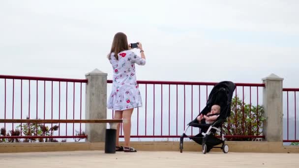 Mamá con un bebé en silla de ruedas en la plataforma de observación toma fotos de hermosas vistas del recuerdo del viaje . — Vídeos de Stock