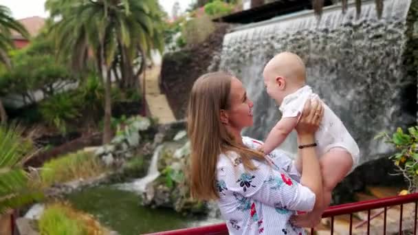 Молодая мама с ребенком, играющим с ним, улыбается и смеется у водопада в парке — стоковое видео