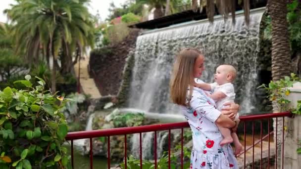 年轻的妈妈抱着一个婴儿, 带着儿子在公园的瀑布旁笑着笑着, 当时她正在前往加那利群岛 — 图库视频影像