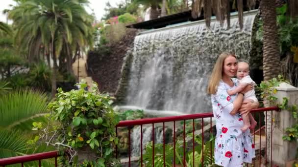 Mutter umarmt Baby und blickt in die Kamera auf dem Hintergrund eines Wasserfalls und Palmen — Stockvideo