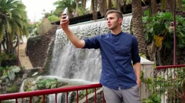 Şelalenin yanında tişörtlü bir adam selfie çeker ve akıllı telefona bir mesaj yazar. Tatilde akıllı bir telefonla uzaktan çalışma.