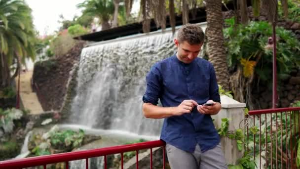 Ein Mann im Hemd, der in der Nähe eines Wasserfalls steht, schreibt eine Nachricht ins Smartphone. Fernarbeit mit dem Smartphone im Urlaub. — Stockvideo