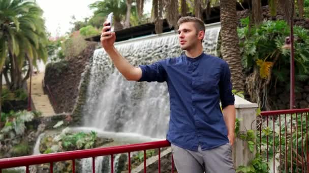 A man in a shirt standing near the waterfall takes a selfie and writes a message in the smartphone. Remote work with a smartphone on vacation. — Stock Video