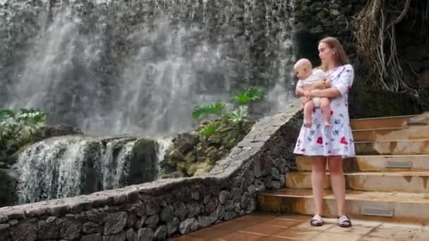 Il bambino seduto alle mani delle madri ridendo e sorridendo guardando la cascata durante un viaggio in famiglia . — Video Stock
