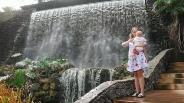 Das Baby sitzt lachend an den Händen seiner Mutter und schaut lächelnd auf den Wasserfall während eines Familienausflugs. — Stockvideo