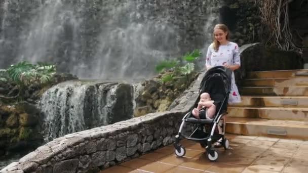 Baby im Rollstuhl lachend und lächelnd den Wasserfall während eines Familienausflugs betrachtend. Mutter geht mit Kind im Kinderwagen. das Konzept einer glücklichen Familie — Stockvideo