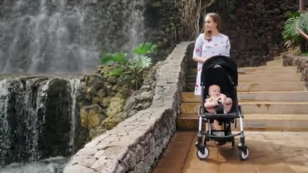 Baby im Rollstuhl lachend und lächelnd den Wasserfall während eines Familienausflugs betrachtend. Mutter geht mit Kind im Kinderwagen. das Konzept einer glücklichen Familie — Stockvideo