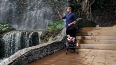 Şelalenin yakınındaki bir tekerlekli sandalyede bir çocukla yürürken bir genç baba sosyal ağlar ve ailesiyle birlikte seyahat ederken Internet yazabilmek için onun Smartphone cep telefonu ile fotoğraf çeker.
